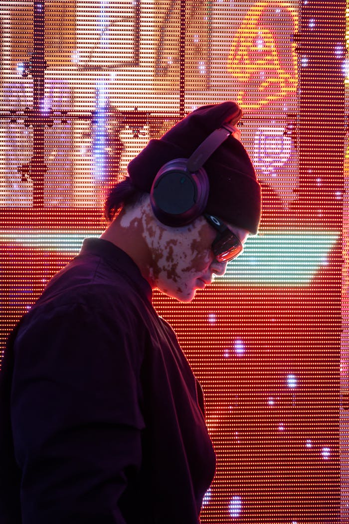 Dj wearing headphones and sunglasses at nightclub with vibrant neon lights and digital screens.